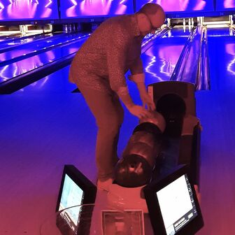 Eine Person beugt sich über die Ballrückgabe einer Bowlingbahn, um eine Kugel auszuwählen. Im Hintergrund ist die Bahn in blau-violettem Neonlicht beleuchtet.