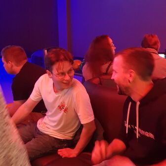 Zwei Männer sitzen lachend auf einer Ledercouch in einer Bowlingbahn mit pink-blauer Neonbeleuchtung. Weitere Personen sitzen im Hintergrund.