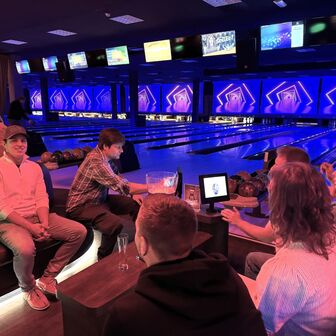 Gruppe von Personen beim Bowling unter blau-violettem Licht, einige sitzen auf Sofas, andere wählen Bowlingkugeln aus.