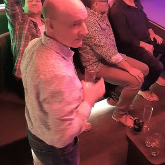 Männer beim Bowlingabend, einer steht mit Getränk, andere sitzen und schauen zu, einer winkt.